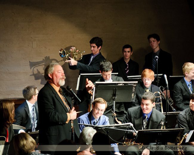Man Speaking On Stage With Musicians Sitting Beside Him